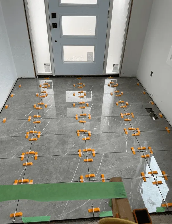 Entryway floor being tiled with large-format polished grey porcelain with white veining, with tile levelling clips in every joint